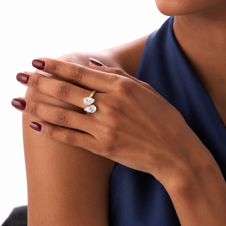 A close-up of a woman's hand wearing a silver ring with two pear-cut clear gemstones, set against a clean white background and a navy blue top.