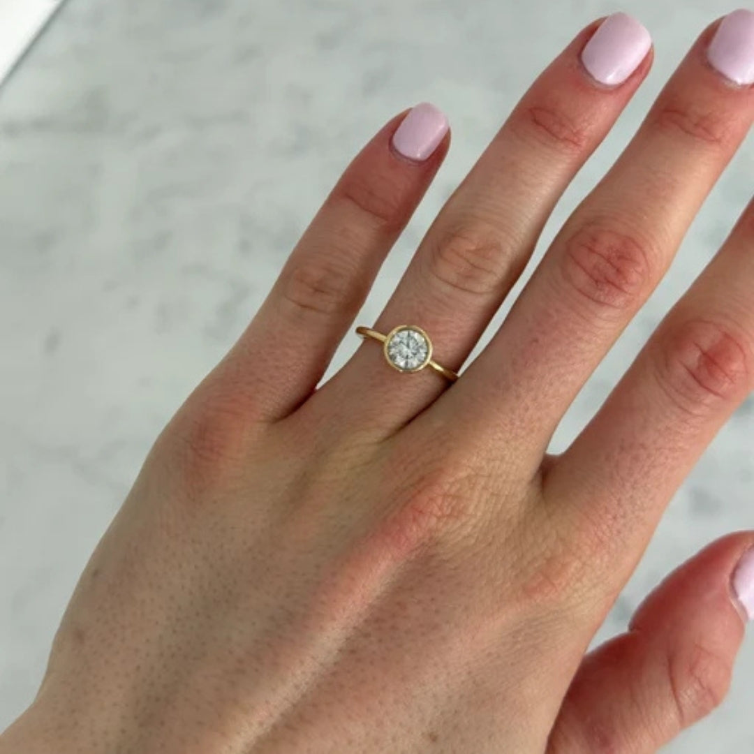 A close-up of a hand wearing a delicate gold ring with a round clear Diamond in a bezel setting, set against a light marble background.