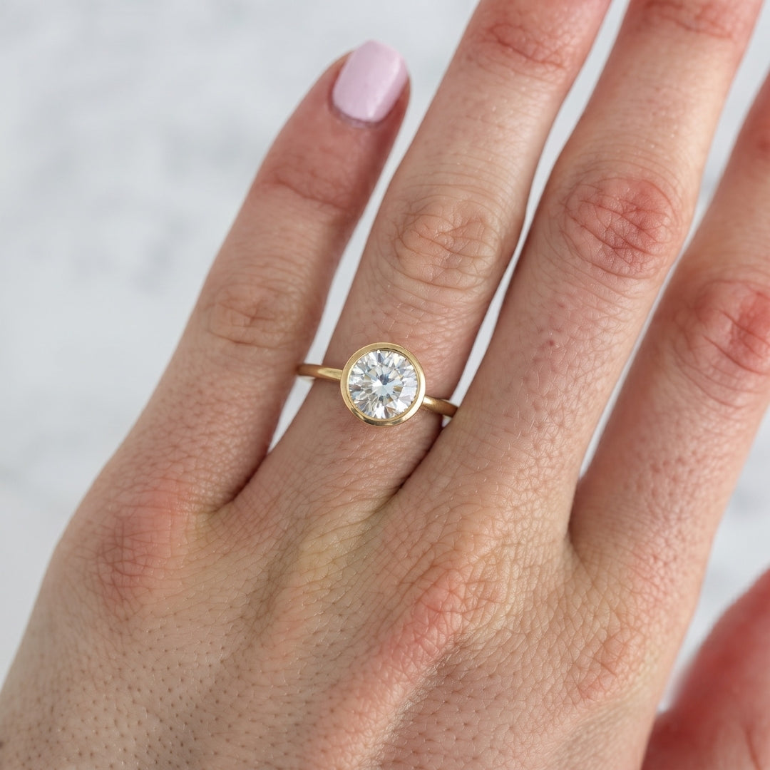 A close-up of a hand wearing a yellow gold ring with a round brilliant-cut diamond. The ring is shown on the middle finger against a soft, light gray marble background.