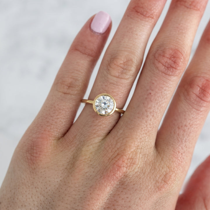 A close-up of a hand wearing a yellow gold ring with a round brilliant-cut diamond. The ring is shown on the middle finger against a soft, light gray marble background.