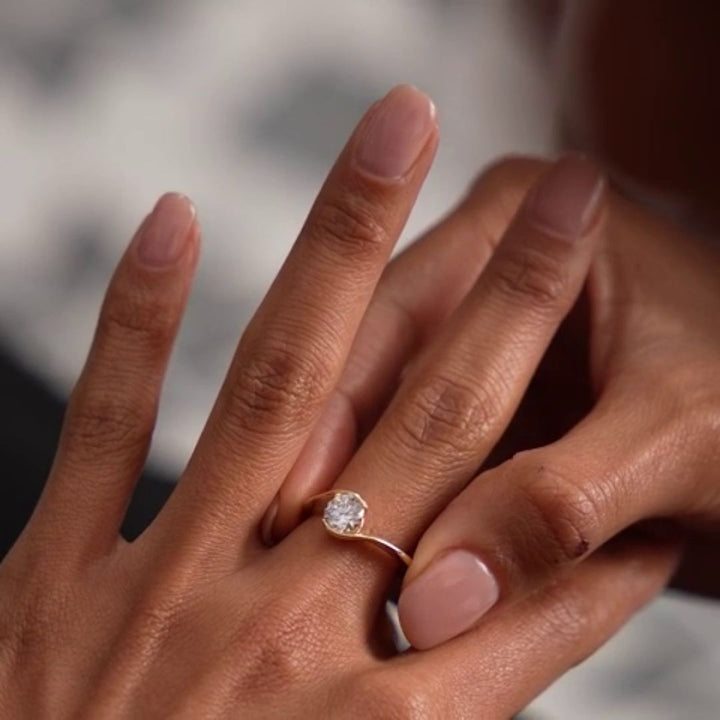 A close-up of a person's hand wearing a delicate gold ring with a round clear diamond, set against a softly blurred background.
