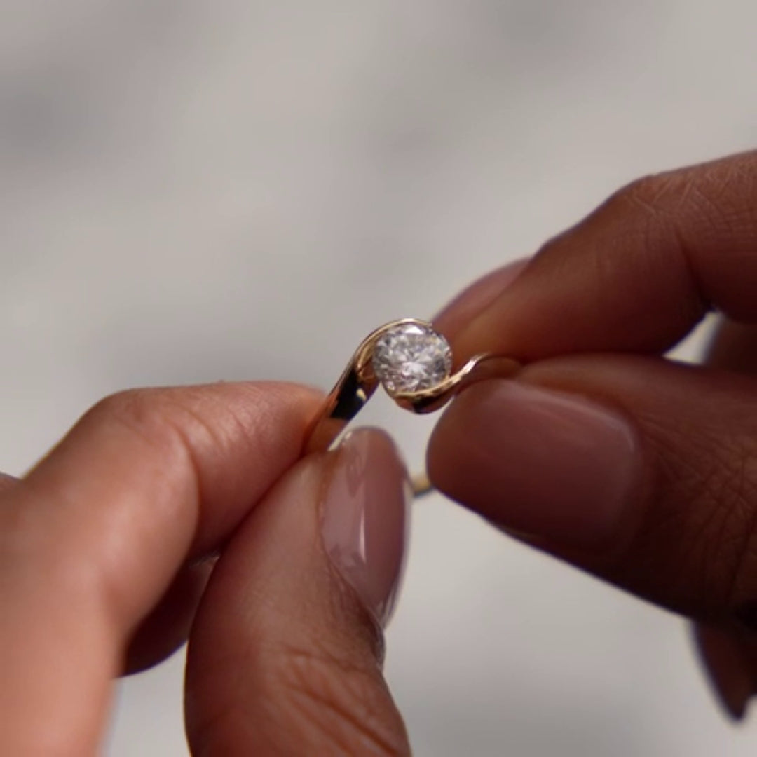 A close-up of a person's hands holding a gold ring with a round clear Diamond against a soft, out-of-focus light gray background.